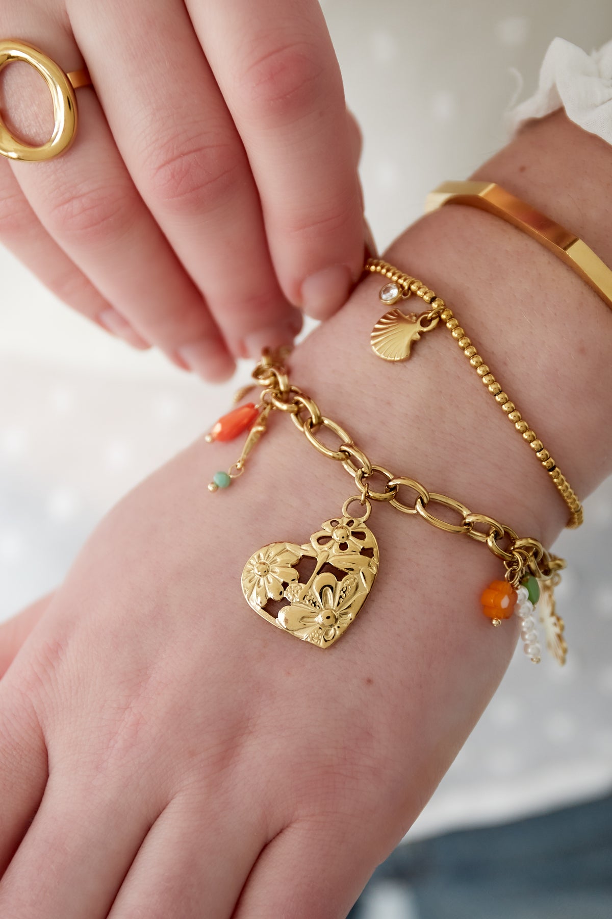 Close-up of wrist wearing gold charm link bracelet alongside two other delicate gold bracelets, showcasing the filigree heart and colourful beads