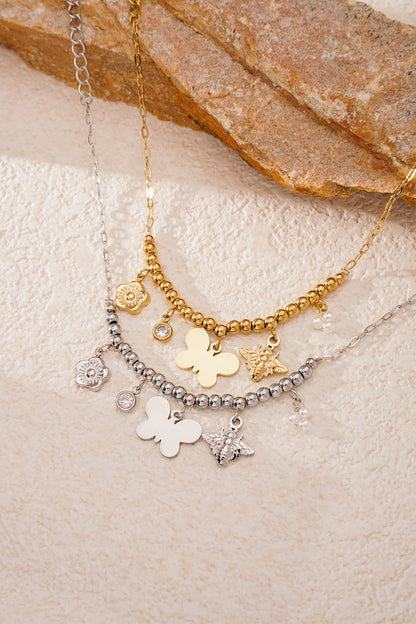 Two charm bracelets, one gold and one silver, both with butterfly, bee, flower, crystal, and pearl charms, placed on a textured stone and cream surface