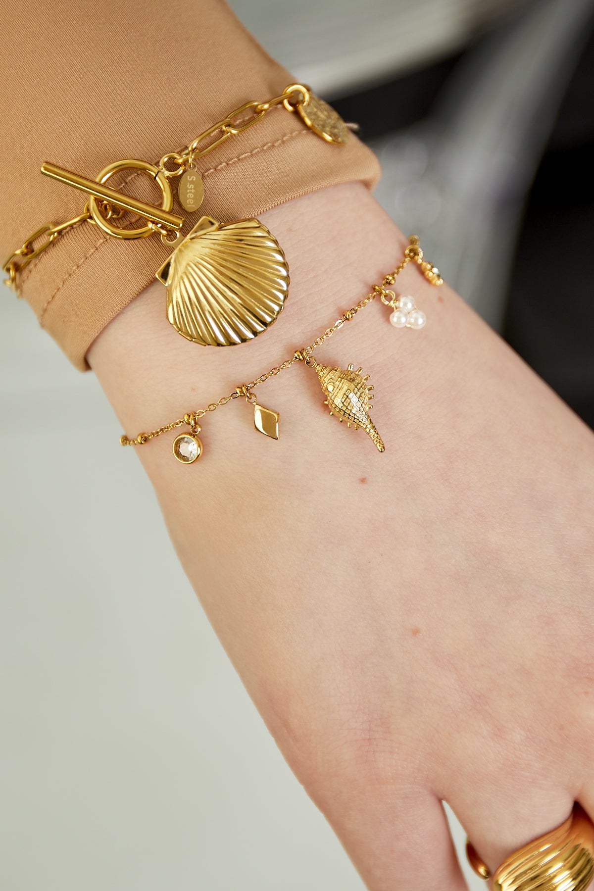 Model wearing gold charm bracelet alongside another gold bracelet with a large shell charm, styled with beige long sleeve and gold rings