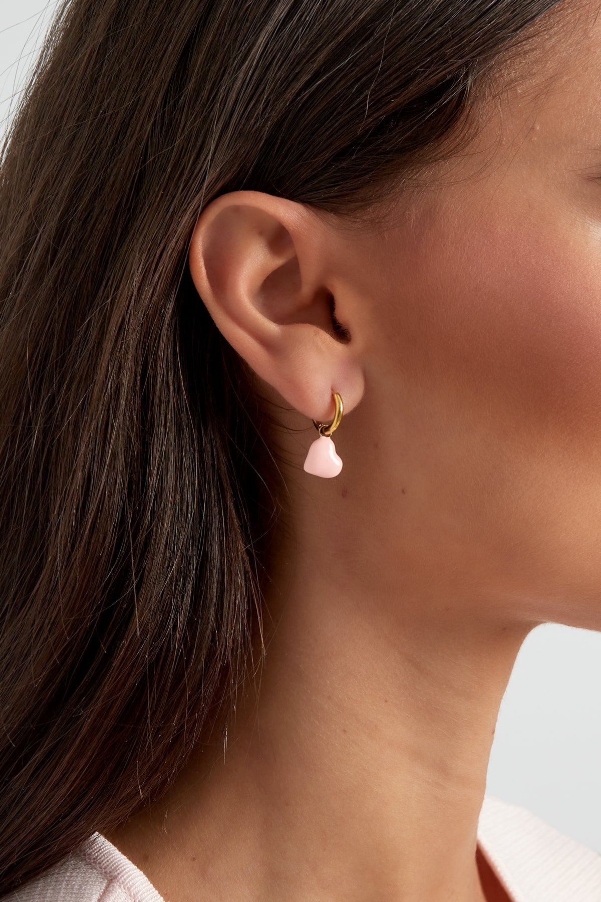 Close-up of woman's ear wearing gold hoop earring with soft pink enamel heart charm