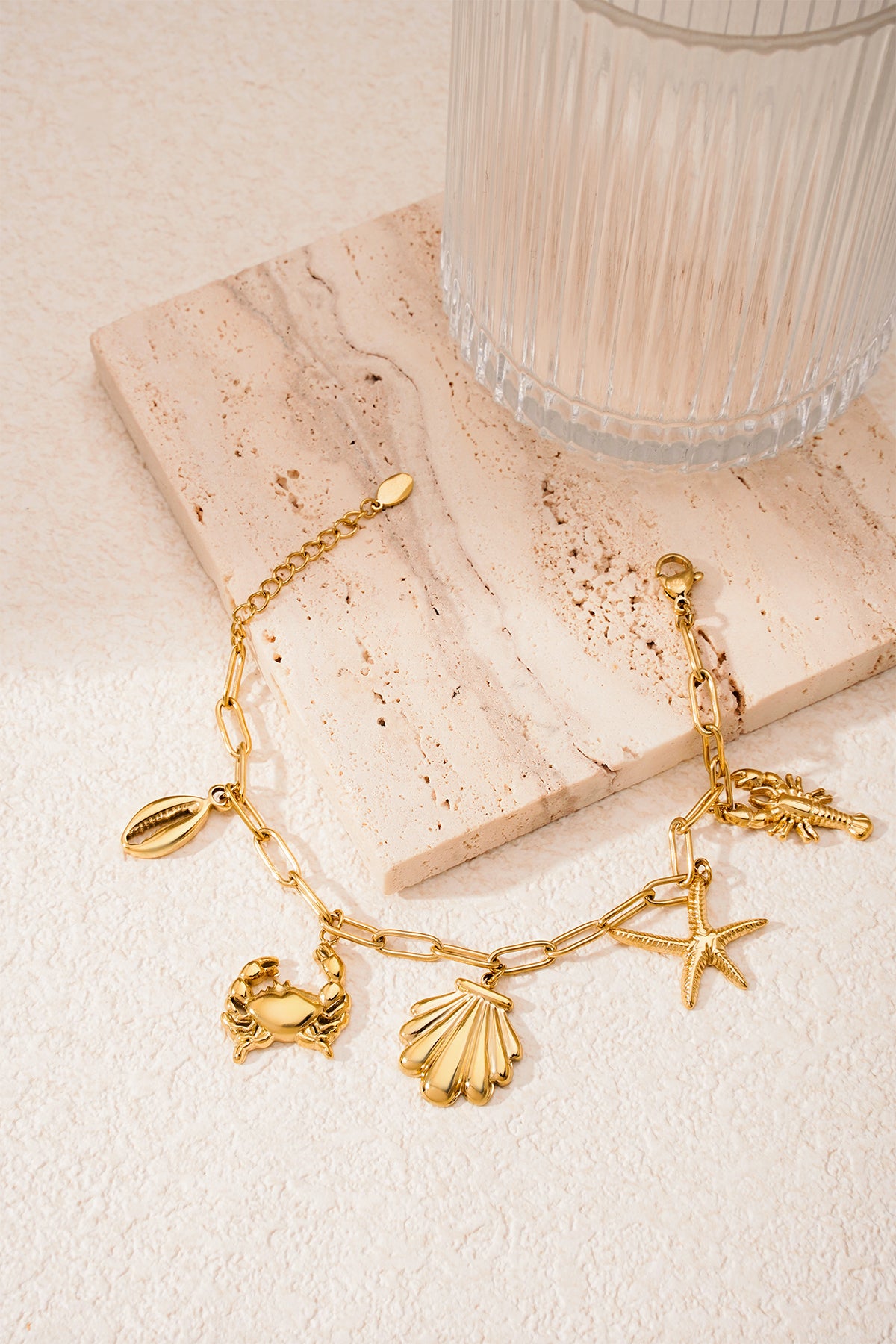 Gold-tone seaside charm bracelet laid on cream textured surface near glass cup
