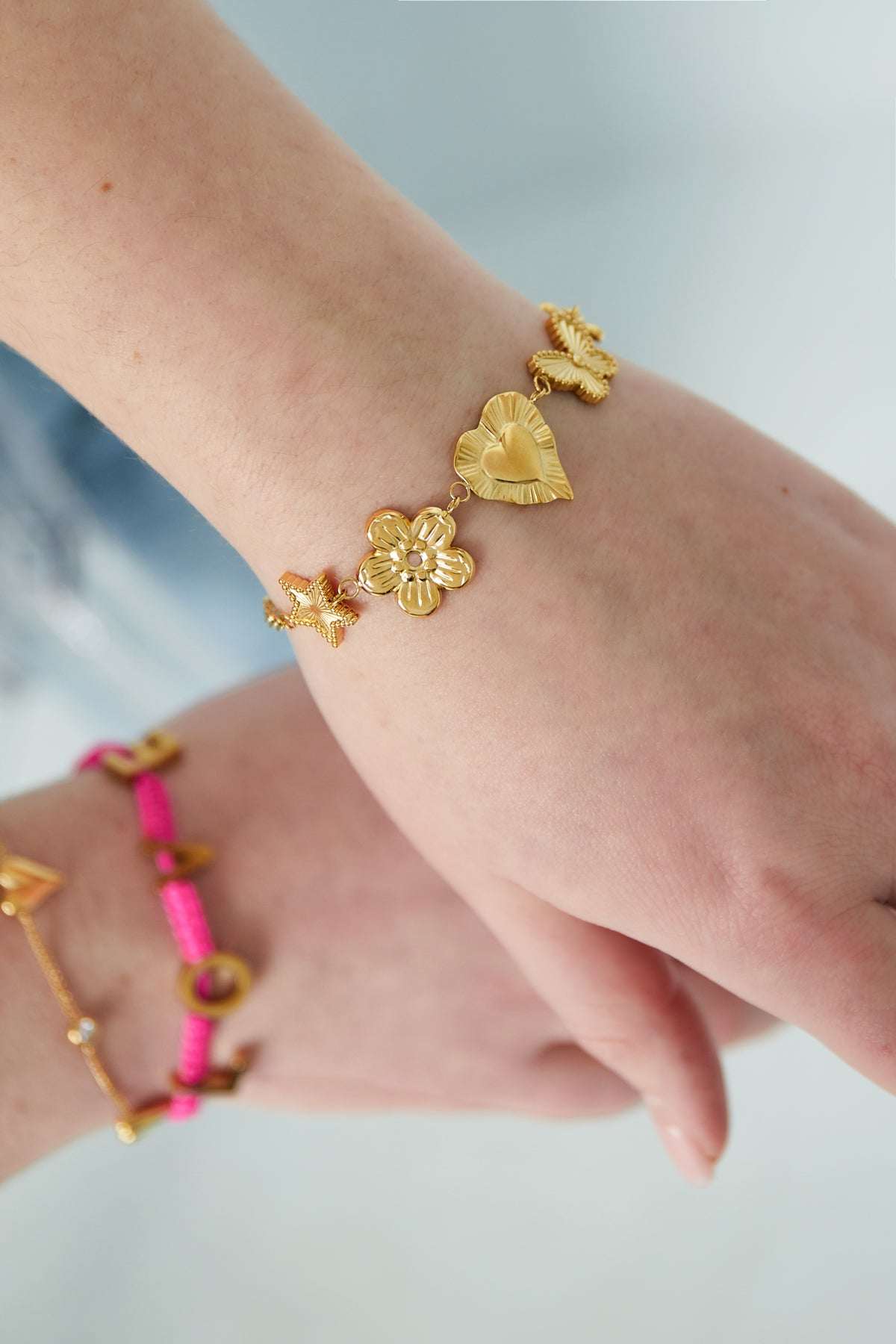 Wrist wearing gold charm bracelet showcasing star, flower, heart, butterfly, and blossom charms styled with pink bracelet