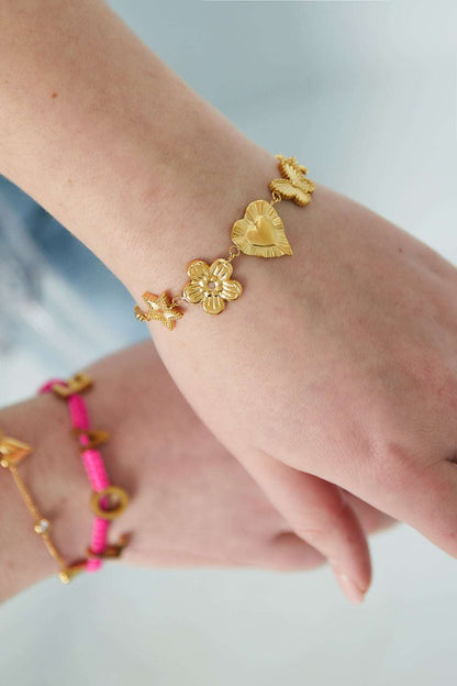 Wrist wearing gold charm bracelet showcasing star, flower, heart, butterfly, and blossom charms styled with pink bracelet