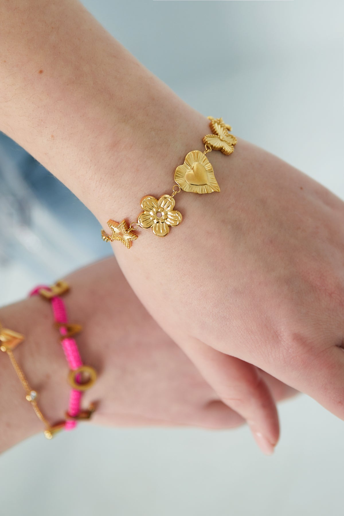 Wrist wearing gold charm bracelet showcasing star, flower, heart, butterfly, and blossom charms styled with pink bracelet