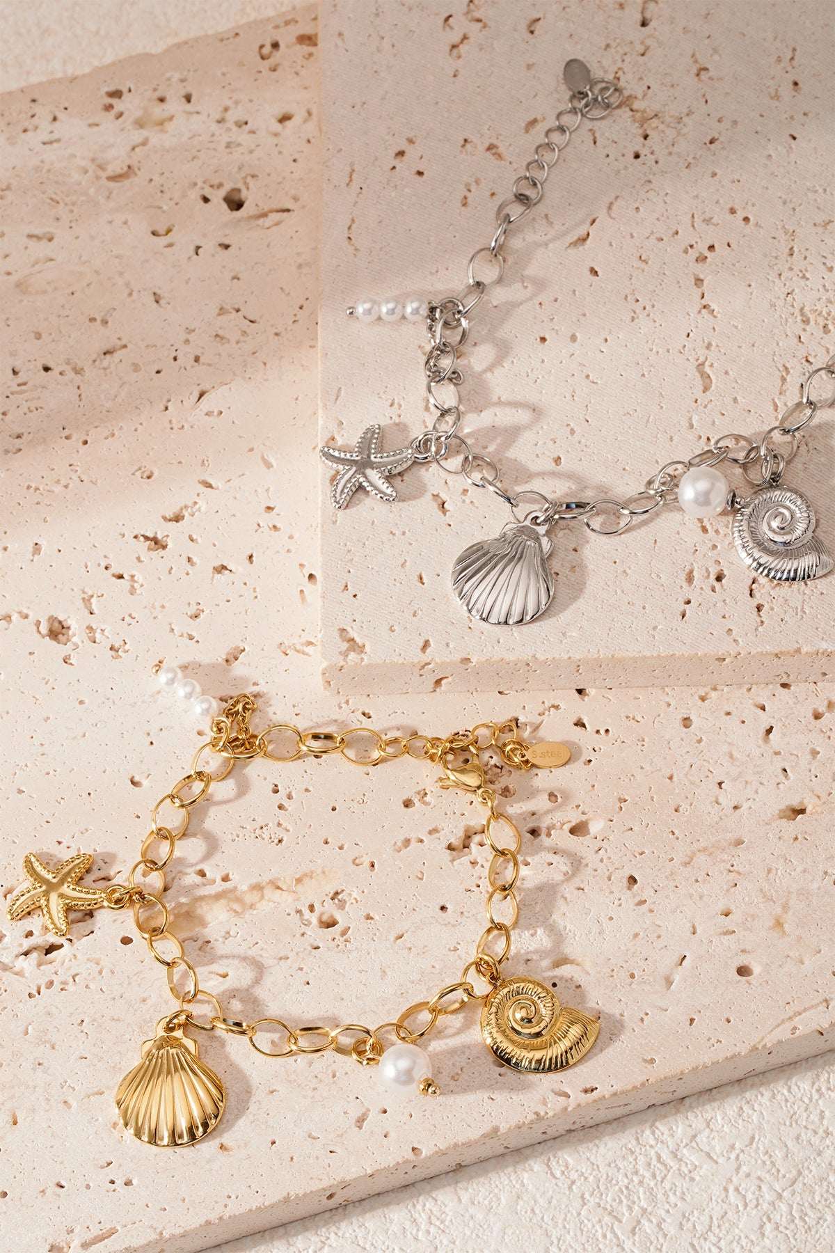 Close-up of gold and silver seaside charm bracelets styled on beige stone tiles with soft natural lighting