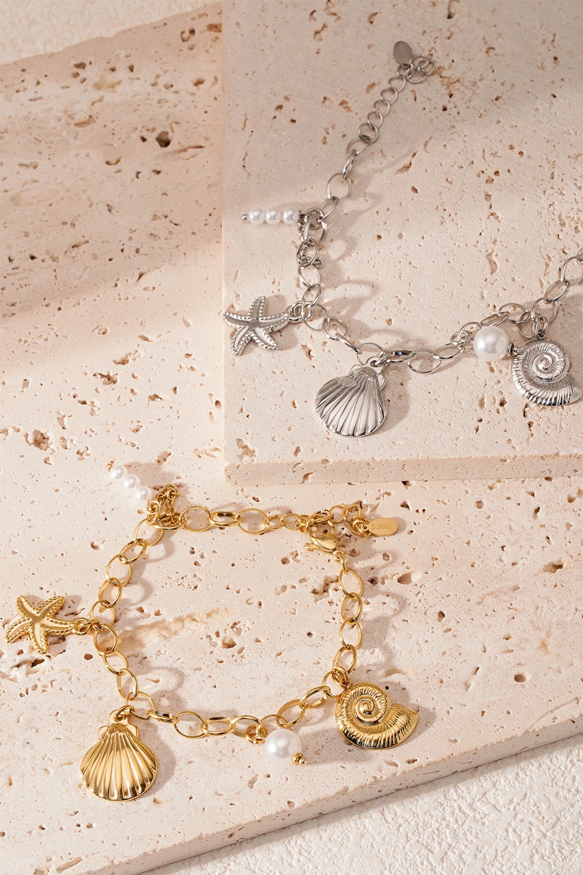 Close-up of gold and silver seaside charm bracelets styled on beige stone tiles with soft natural lighting