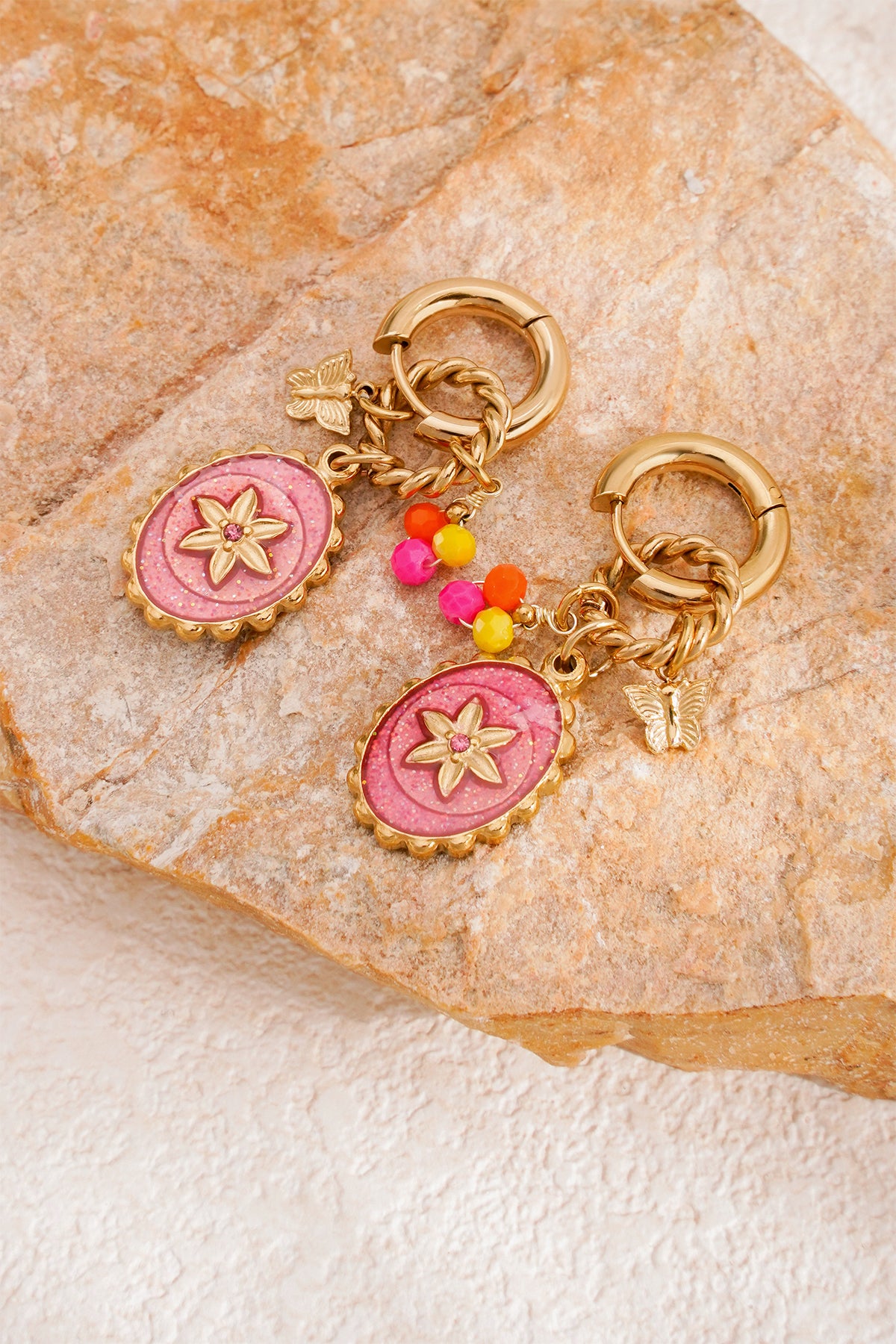 Pair of gold hoop earrings with pink flower pendants and colorful beads resting on textured tan stone surface