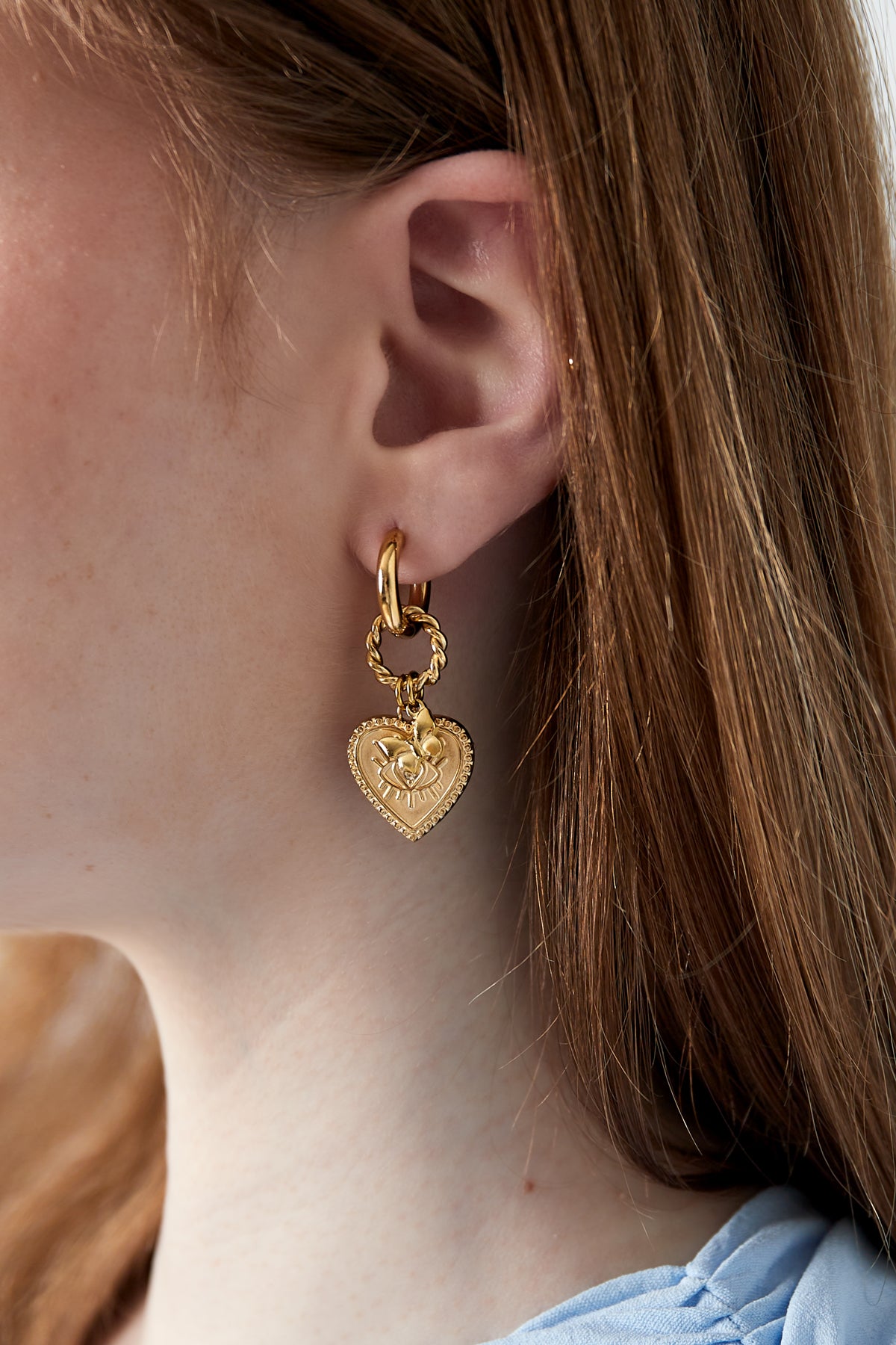 Gold heart charm hoop earring worn on ear with soft brown hair and light blue top