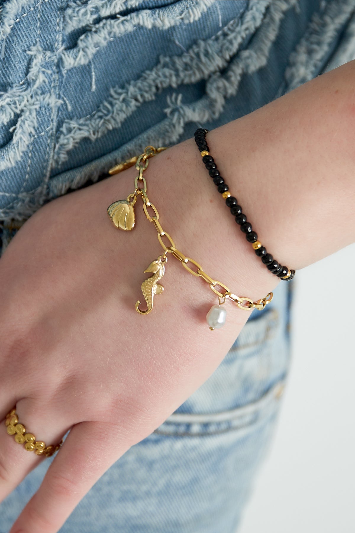 Close-up of wrist wearing a black and gold beaded bracelet paired with a gold charm bracelet featuring seashell, seahorse, and pearl charms over denim jeans
