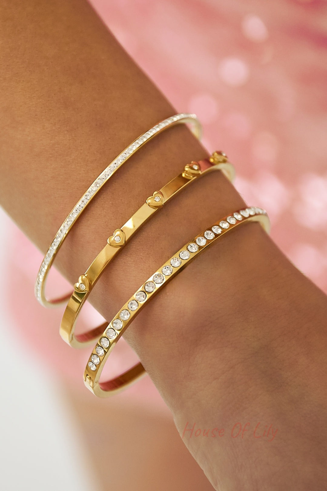 Close-up of a wrist wearing three gold-tone bangles: heart-accented, rhinestone-lined, and plain minimalist styles against a pink background