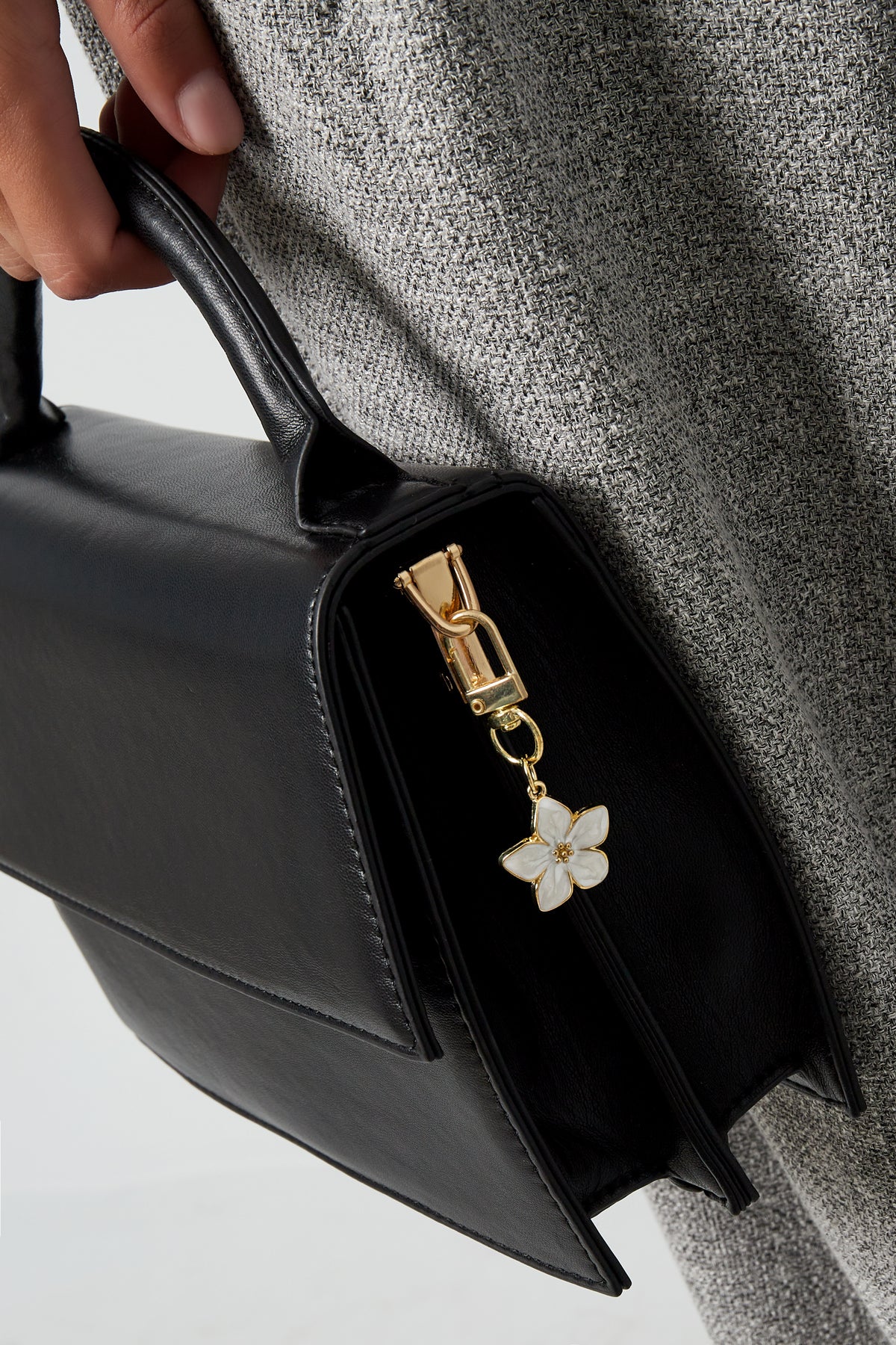 Close-up of a black handbag held by a person wearing gray tweed showing a white enamel blossom flower charm keyring attached to zipper