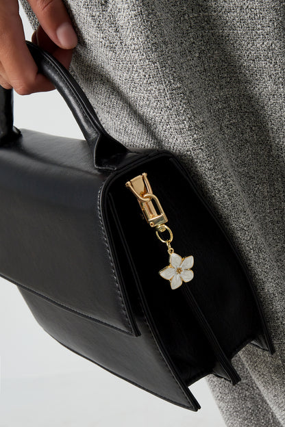 Close-up of a black handbag held by a person wearing gray tweed showing a white enamel blossom flower charm keyring attached to zipper