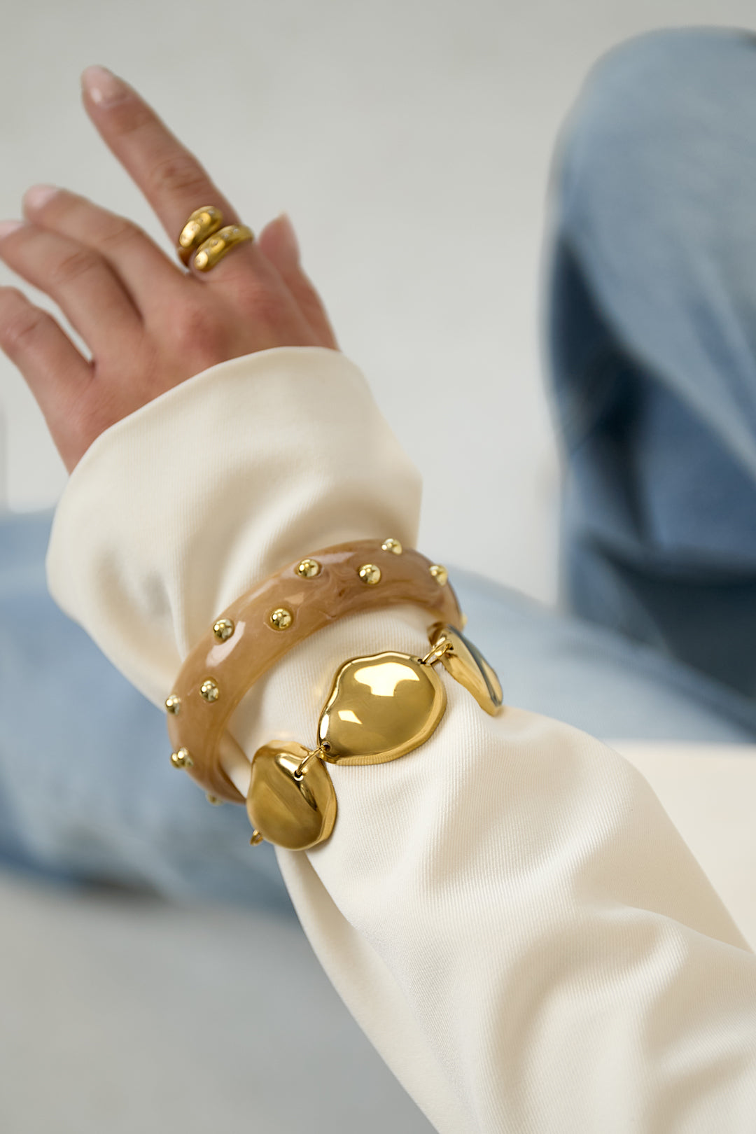 Close-up view of gold chunky bracelet paired with beige studded resin bangle worn on wrist with cream blouse and blue jeans