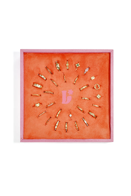 Top view of coral-orange velvet display holding 28 unique gold-tone rings arranged in a circle with a pink border