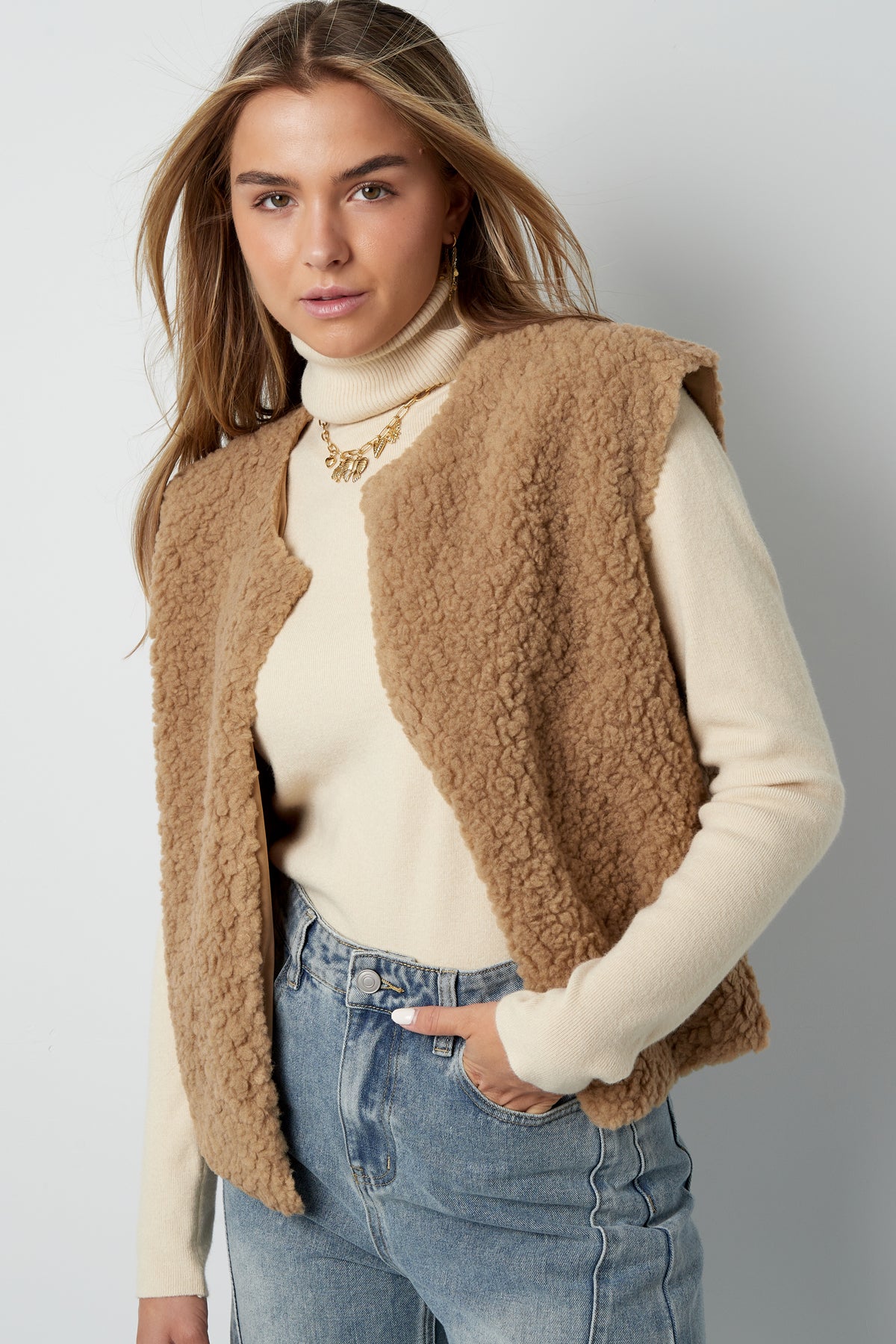 Model styled in camel sherpa vest layered over cream turtleneck sweater and light washed jeans