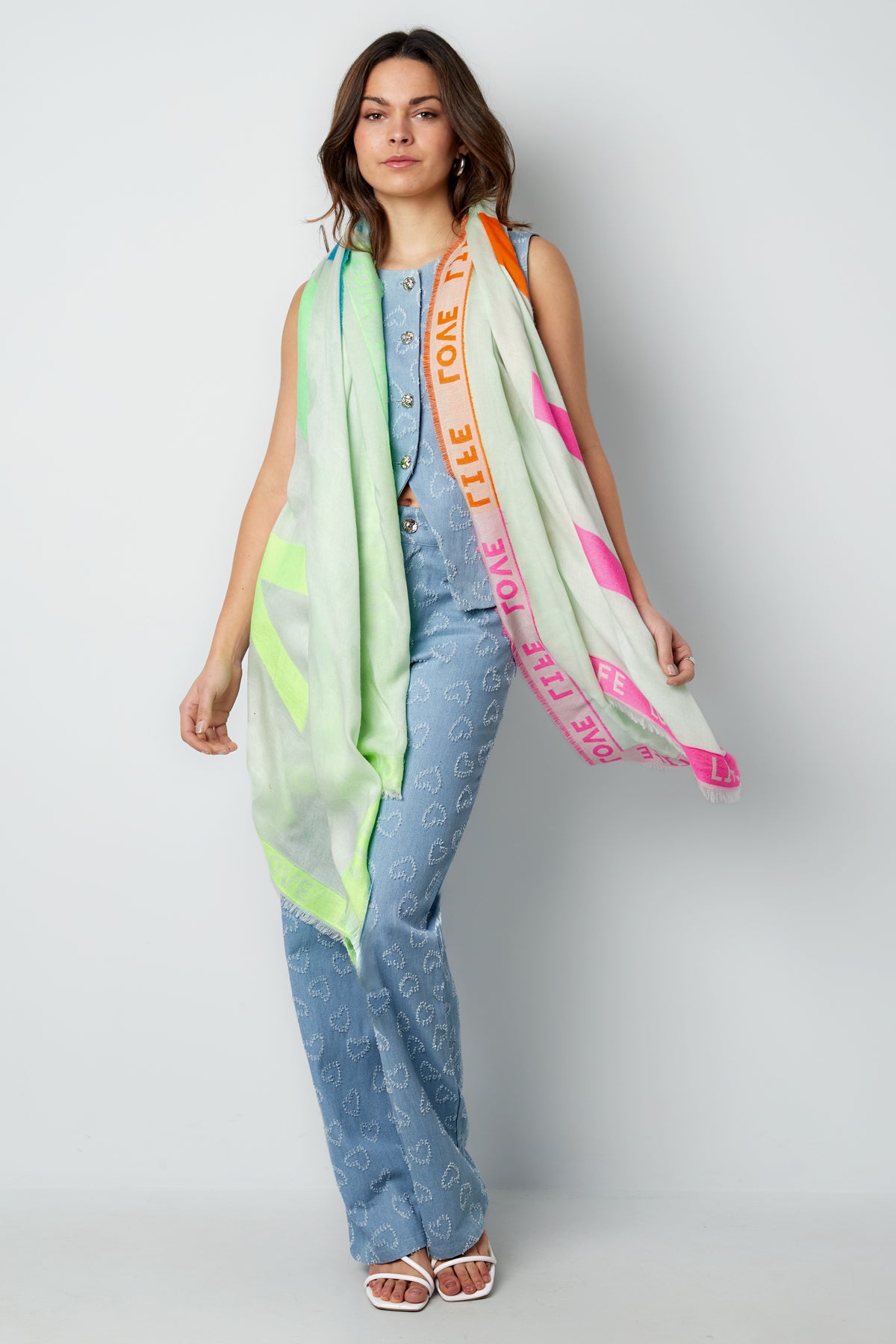 Model wearing colorful LIFE LOVE scarf draped around shoulders, styled with blue patterned outfit and white sandals