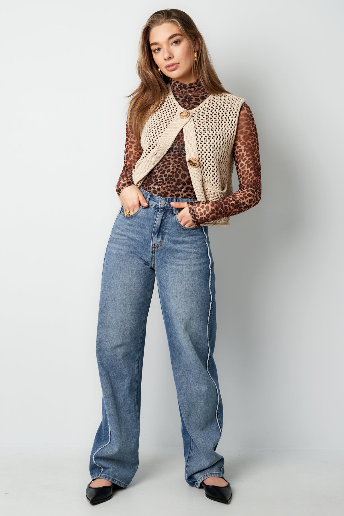 Close up side view of model in beige chunky knit vest with leopard print top and jeans