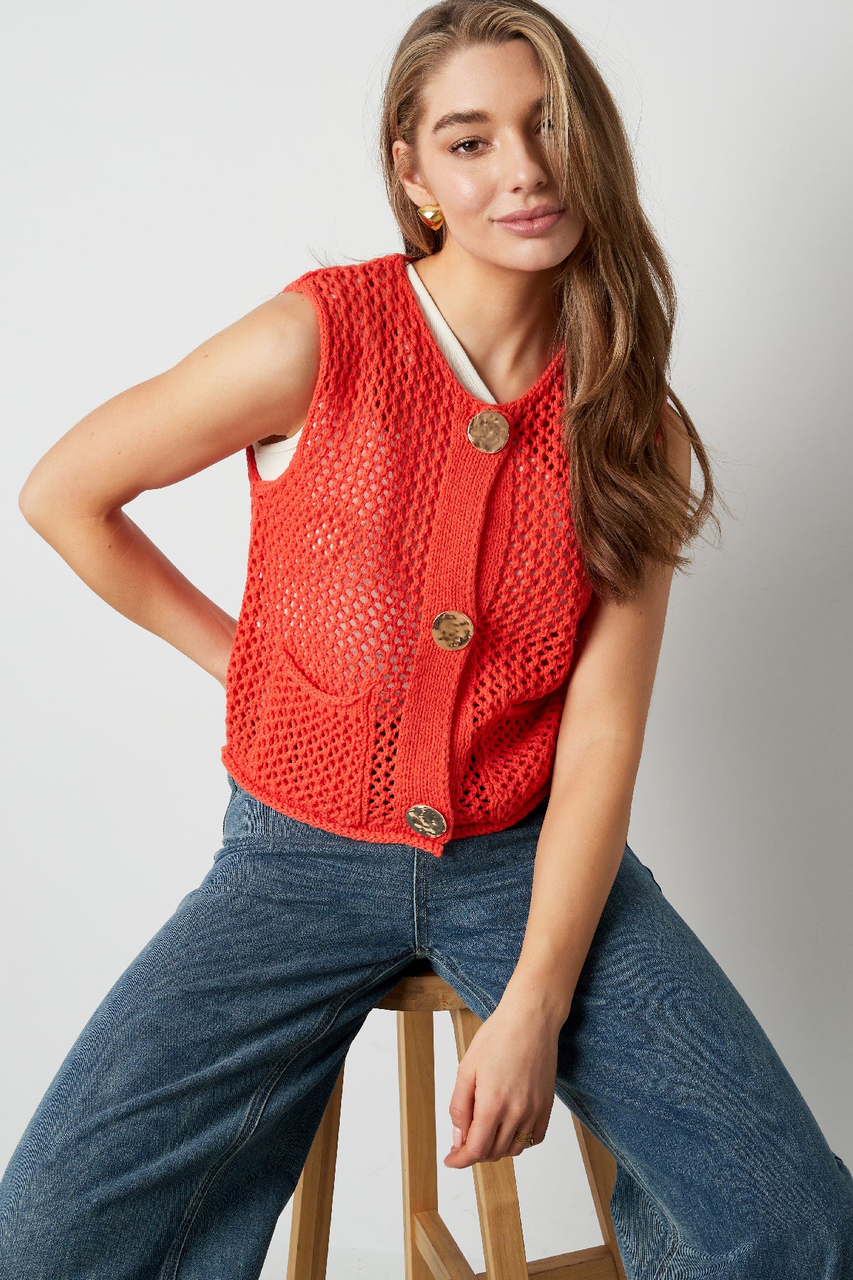Model wearing bright red chunky knit buttoned vest paired with jeans sitting on stool
