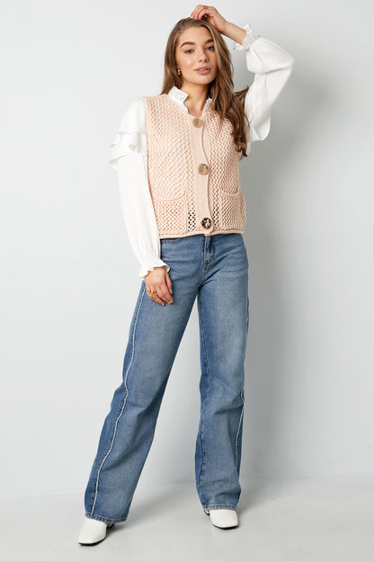 Full-length photo of model in beige knit vest layered over ruffled white blouse and wide-leg jeans