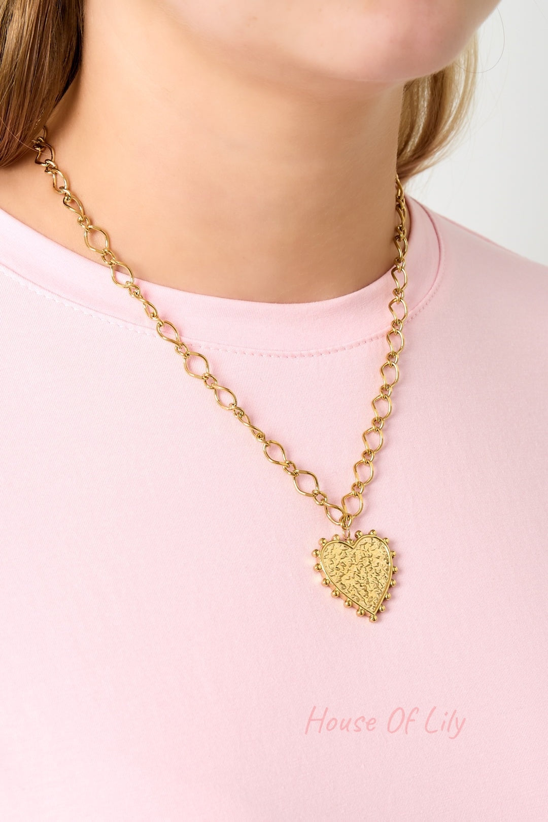 Gold-tone heart pendant necklace worn on a model's neck over a pink shirt, showing the necklace's medium length and detailed design