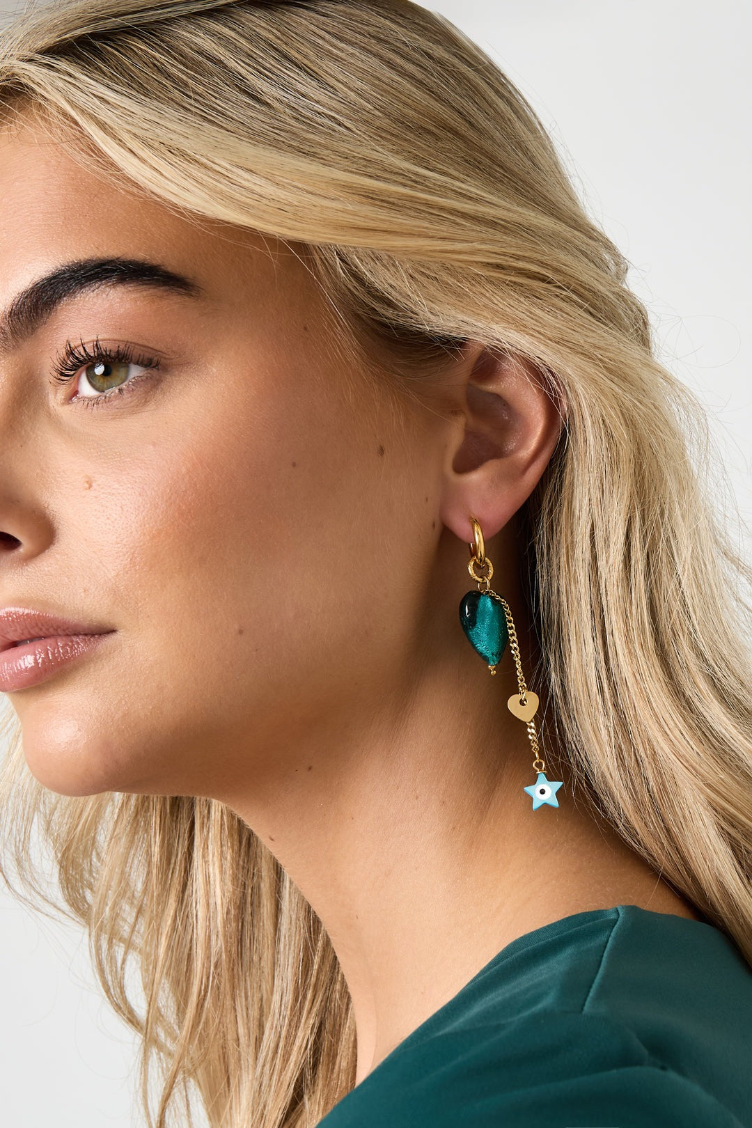 Side view of a woman wearing gold-tone drop earring with emerald-green heart, small gold heart charm, and blue star pendant dangling