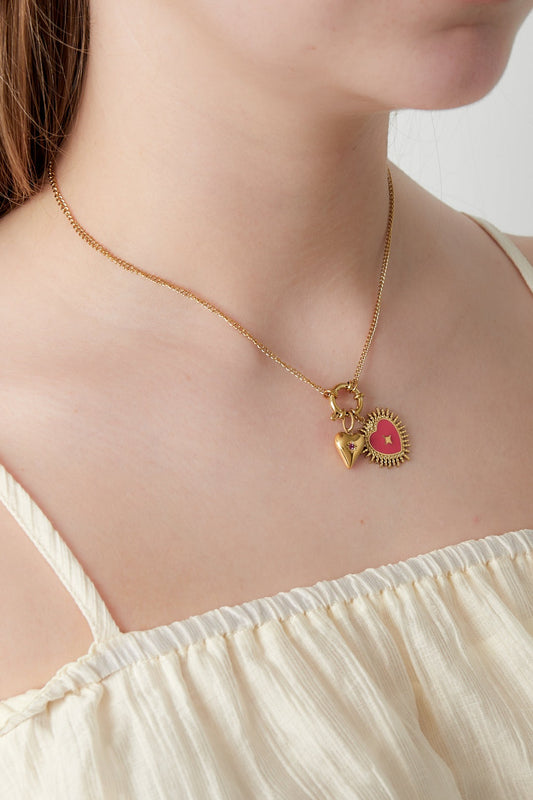 Close-up of gold pendant necklace featuring red sunburst heart and small gold heart charm worn with cream top
