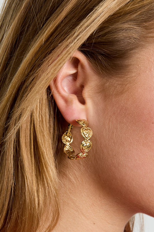 Gold-tone hoop earrings featuring a linked chain design of embossed, rounded hearts worn on a model's ear.