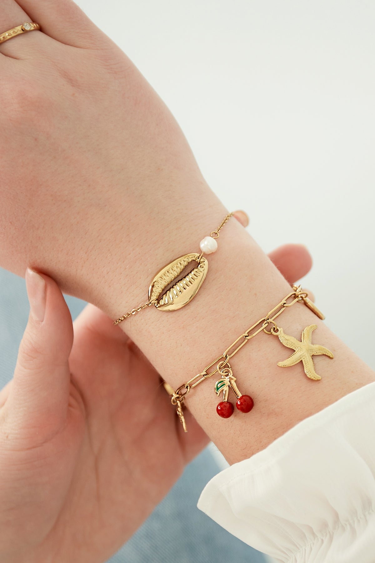 Close-up of a wrist wearing the gold charm bracelet alongside a delicate pearl and shell bracelet, showcasing the red cherry and starfish charms