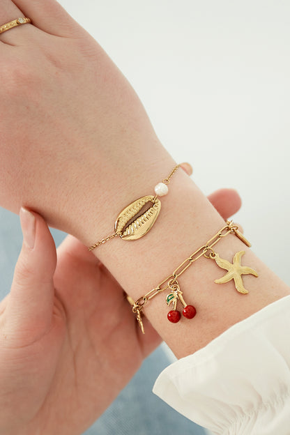 Close-up of a wrist wearing the gold charm bracelet alongside a delicate pearl and shell bracelet, showcasing the red cherry and starfish charms