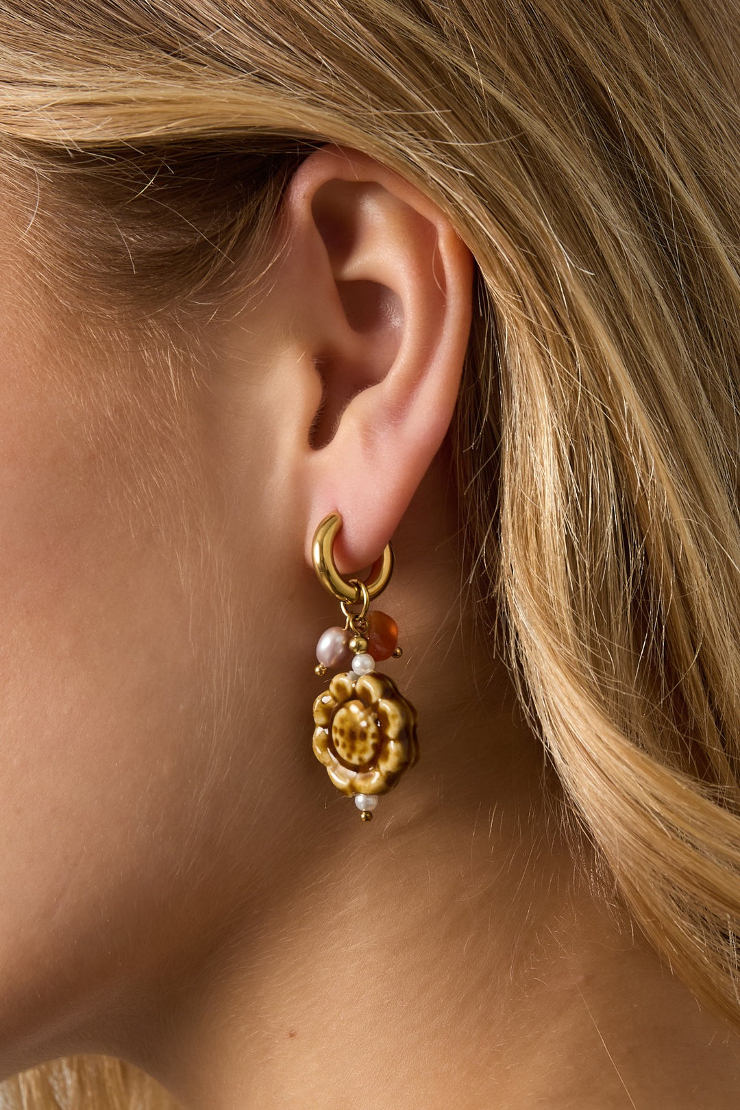 Close-up side profile of woman wearing one artisanal floral charm earring highlighting textured ceramic flower, pearl, coral beads, and gold hoop closure