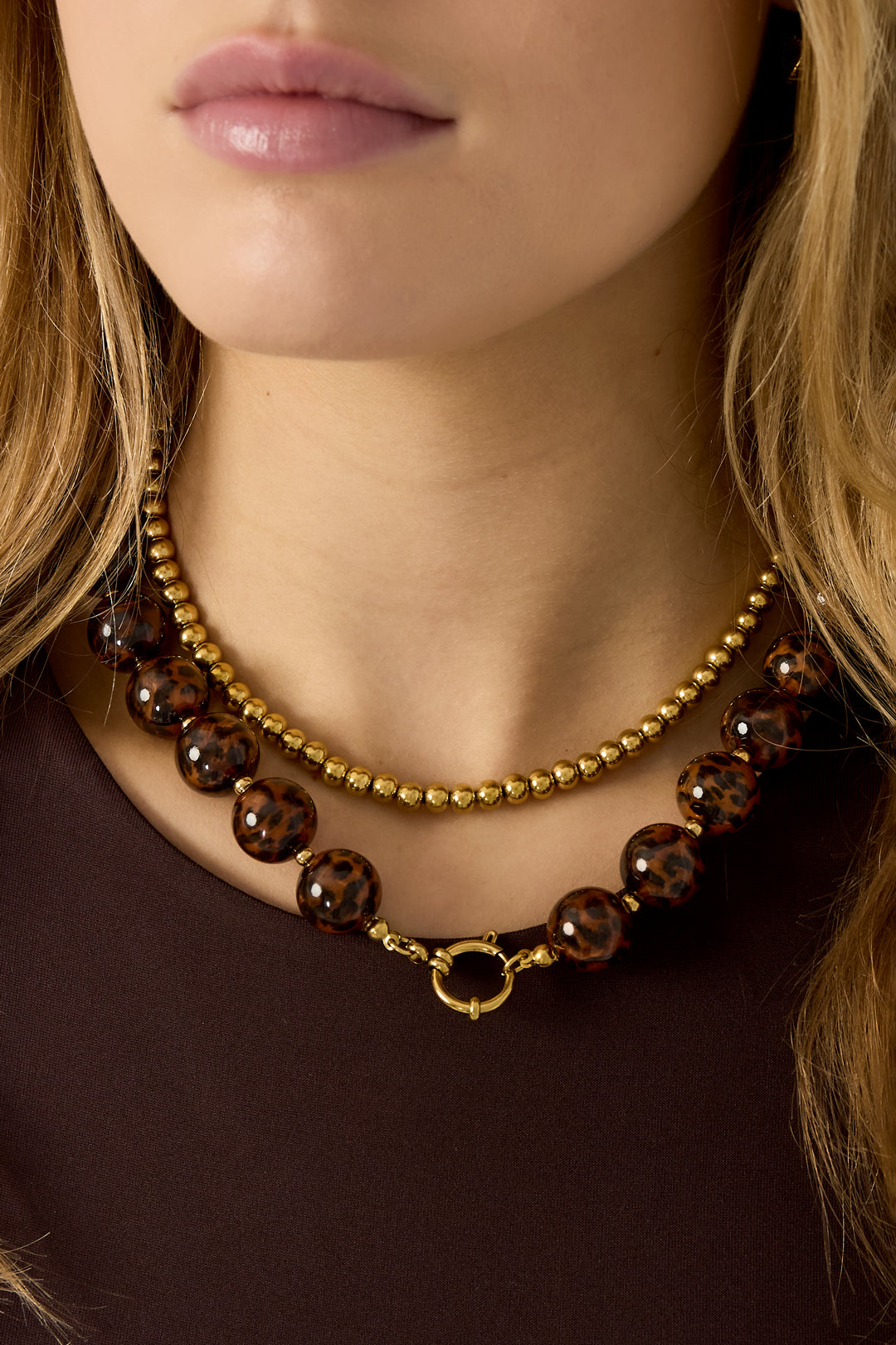 Model wearing layered necklaces featuring shiny gold beads and a leopard print beaded necklace with gold clasp over a brown top