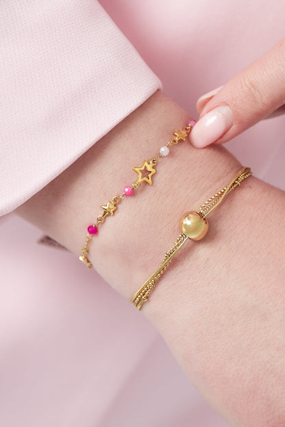 Close-up of wrist wearing the gold star charm bracelet with pink beads alongside a second gold chain bracelet, set against soft pink background