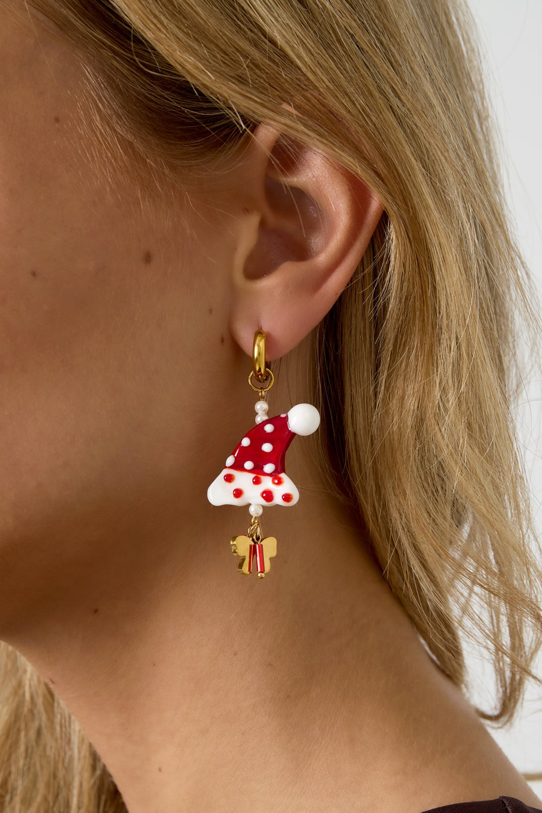 Close-up side view of a woman wearing a festive Santa hat drop earring with red, white, pearl, and gold details.