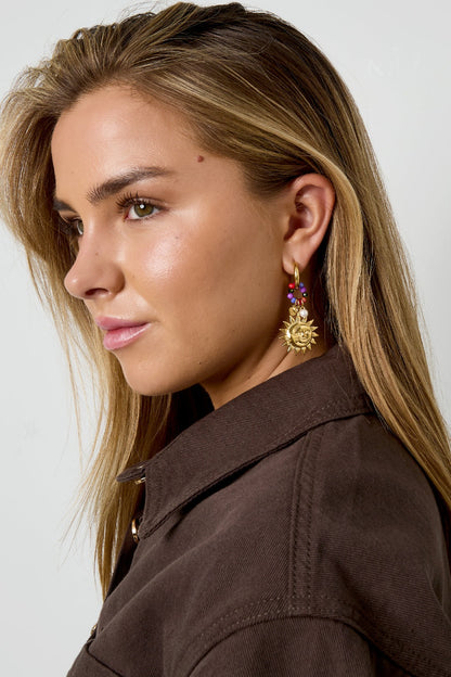 Close-up side view of woman wearing golden sun charm earrings with purple, pink, and multicolor beads, styling with brown jacket