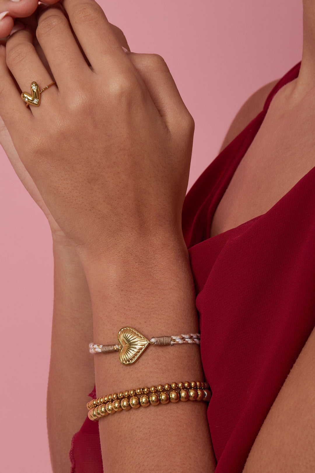 Woman wearing a beige and gold heart charm bracelet with gold bead bracelets and a red top, displayed on arm