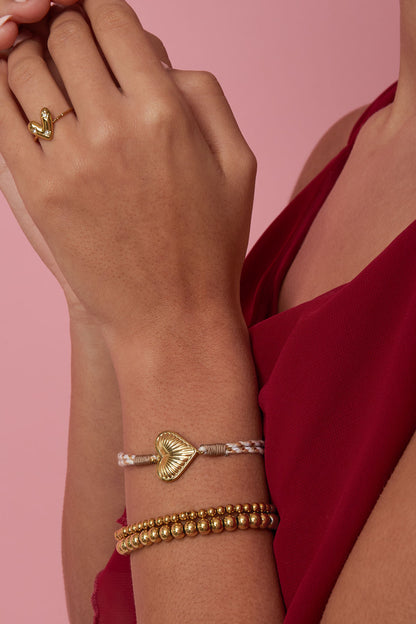 Woman wearing a beige and gold heart charm bracelet with gold bead bracelets and a red top, displayed on arm