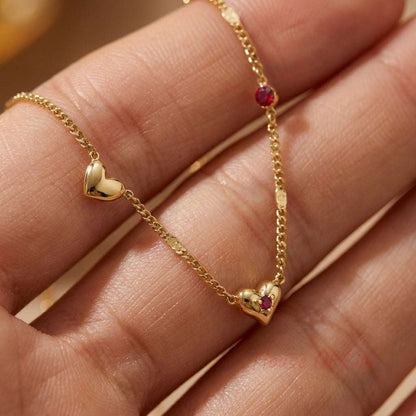 Gold-tone chain necklace with two polished gold hearts, one accented with a small red stone, held in a hand.
