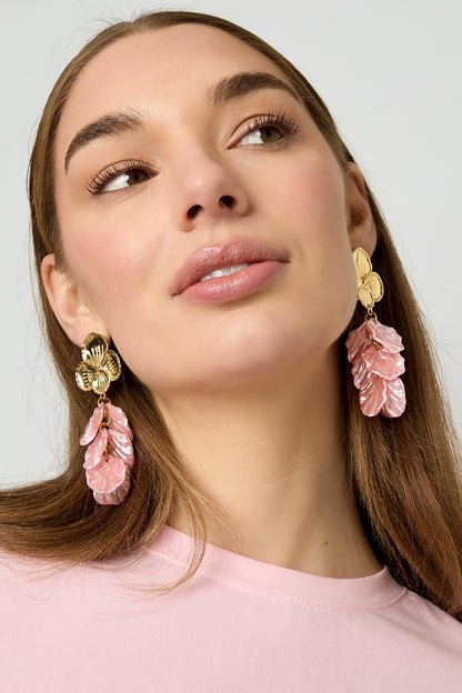 Close-up of a woman wearing gold floral drop earrings with pink layered petals, styled with light pink top