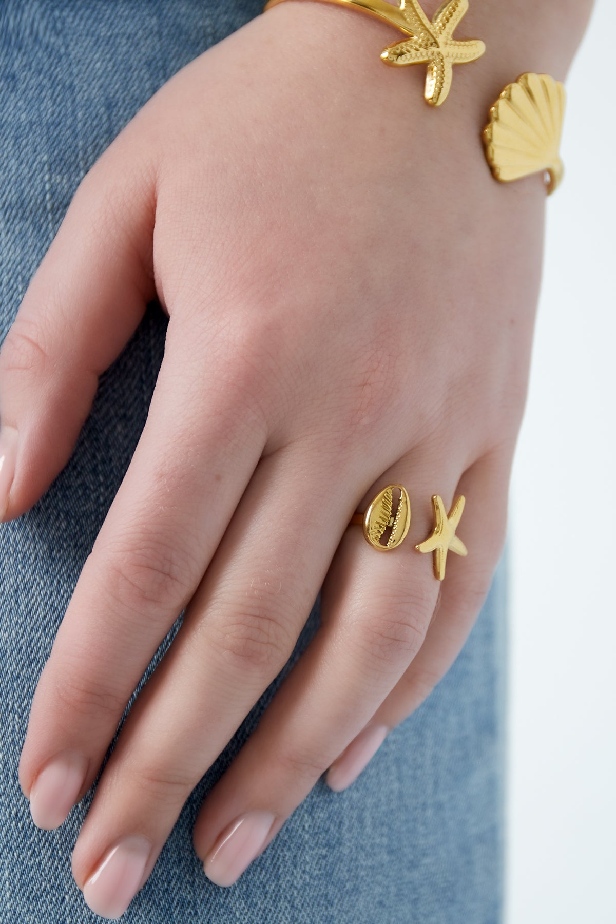 Close-up of hand wearing a gold-tone starfish and seashell open ring with blue denim background
