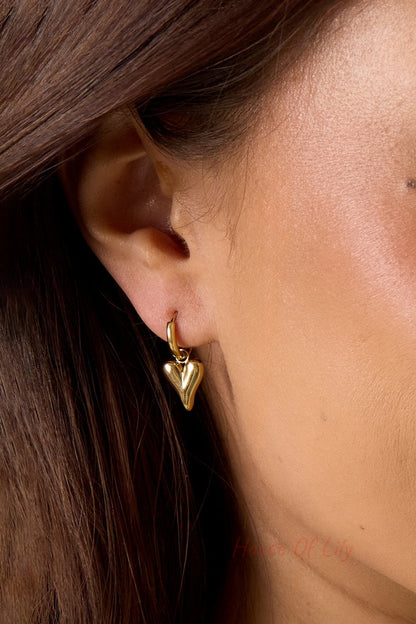 Main Character Heart Drops: A polished gold hoop earring with a dangling, puffy heart charm worn on a earlobe.