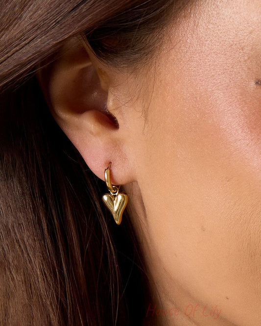Main Character Heart Drops: A polished gold hoop earring with a dangling, puffy heart charm worn on a earlobe.