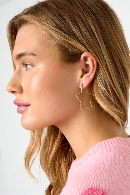 Side profile of woman wearing gold star-shaped textured hoop earrings with loose blonde hair and pink sweater