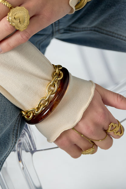 Woman's wrist wearing gold twisted link bracelet layered with a brown resin bangle and rings