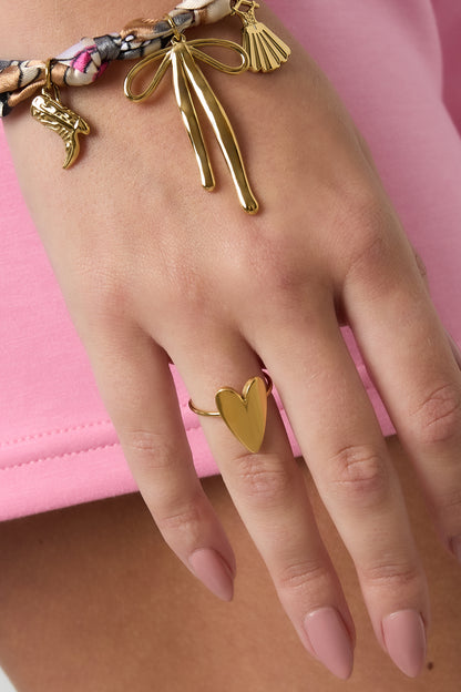 Gold heart ring worn on finger paired with a playful charm bracelet featuring small golden trinkets and a pink outfit
