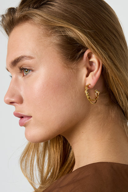 Petit Coeur Hoops: Gold-toned hoop earrings with a small, delicate intertwined heart motif worn by a woman.