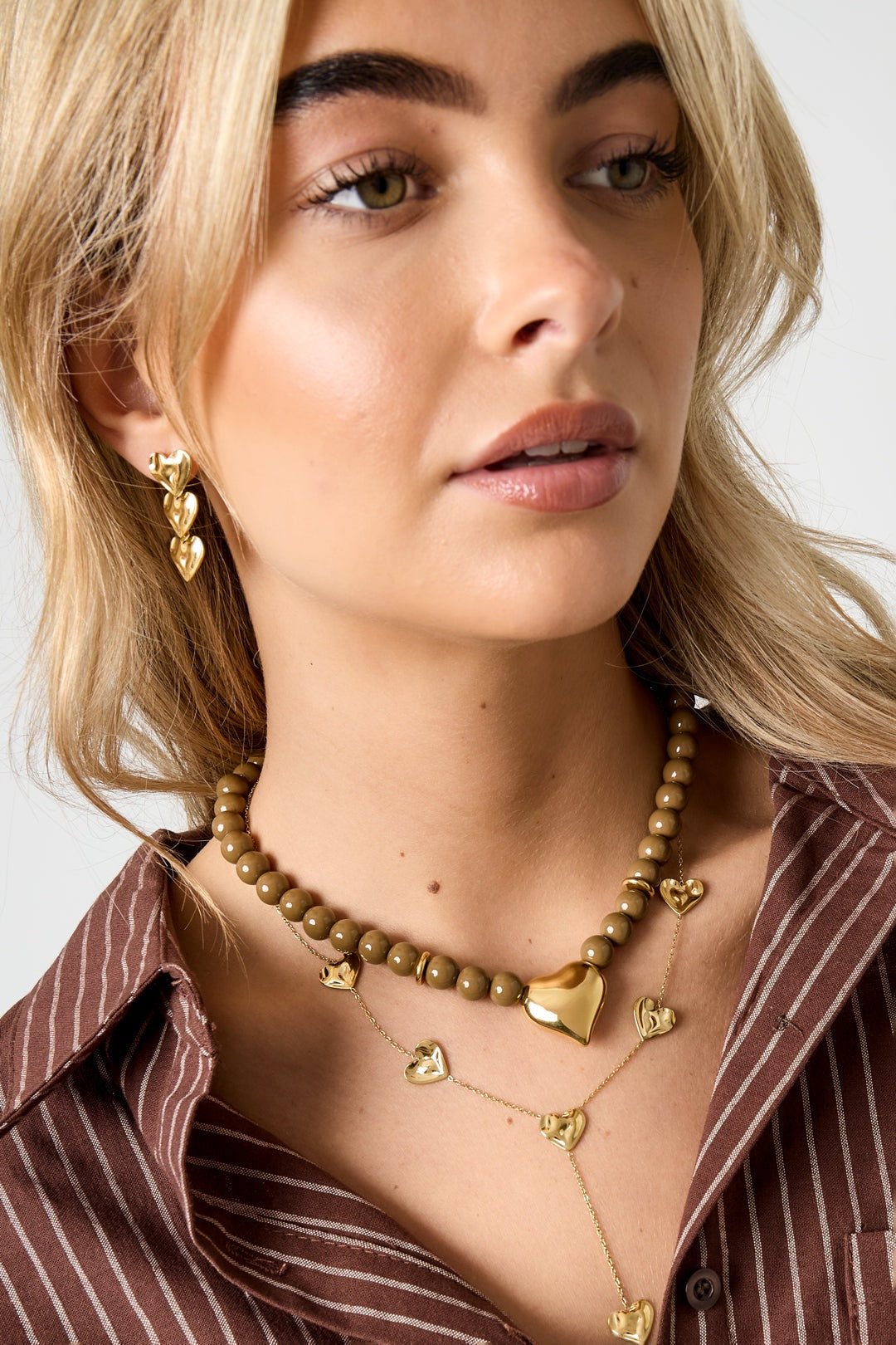 Model wearing olive-toned beaded necklace with gold heart pendant and matching layered heart necklaces along with gold heart earrings against brown striped shirt