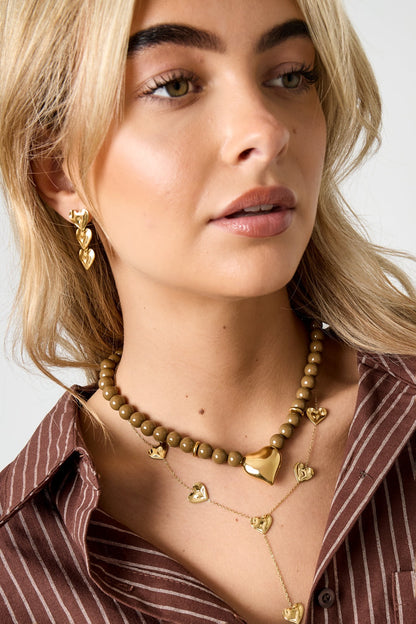 Model wearing olive-toned beaded necklace with gold heart pendant and matching layered heart necklaces along with gold heart earrings against brown striped shirt