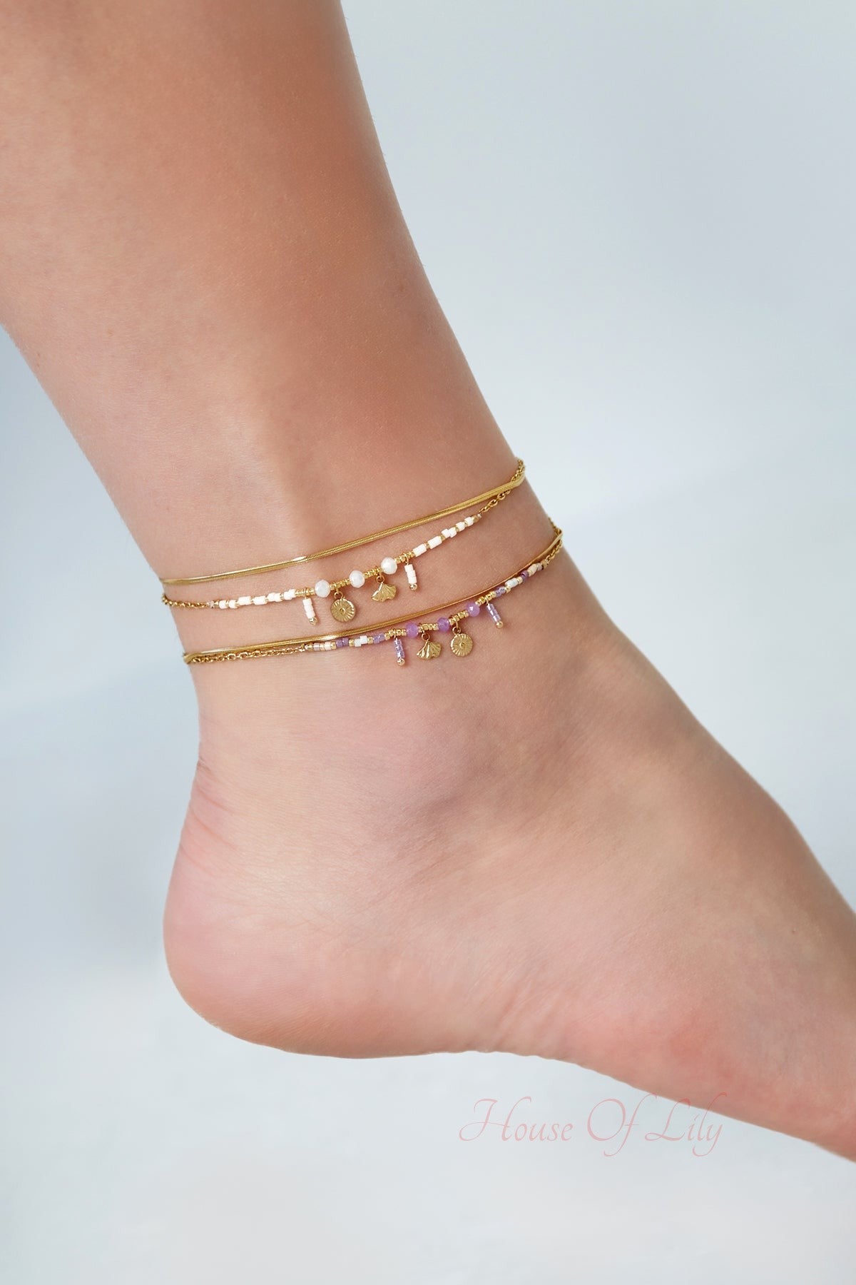 Close-up view of anklets with gold chains, pastel beads, and charms worn on ankle against light background