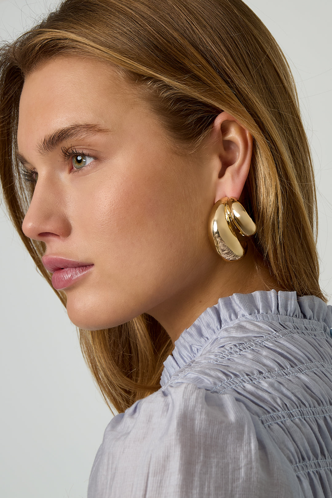 Angled side view of woman showcasing chunky gold hoop earrings with transparent post stoppers, wearing a light blue ruffled blouse