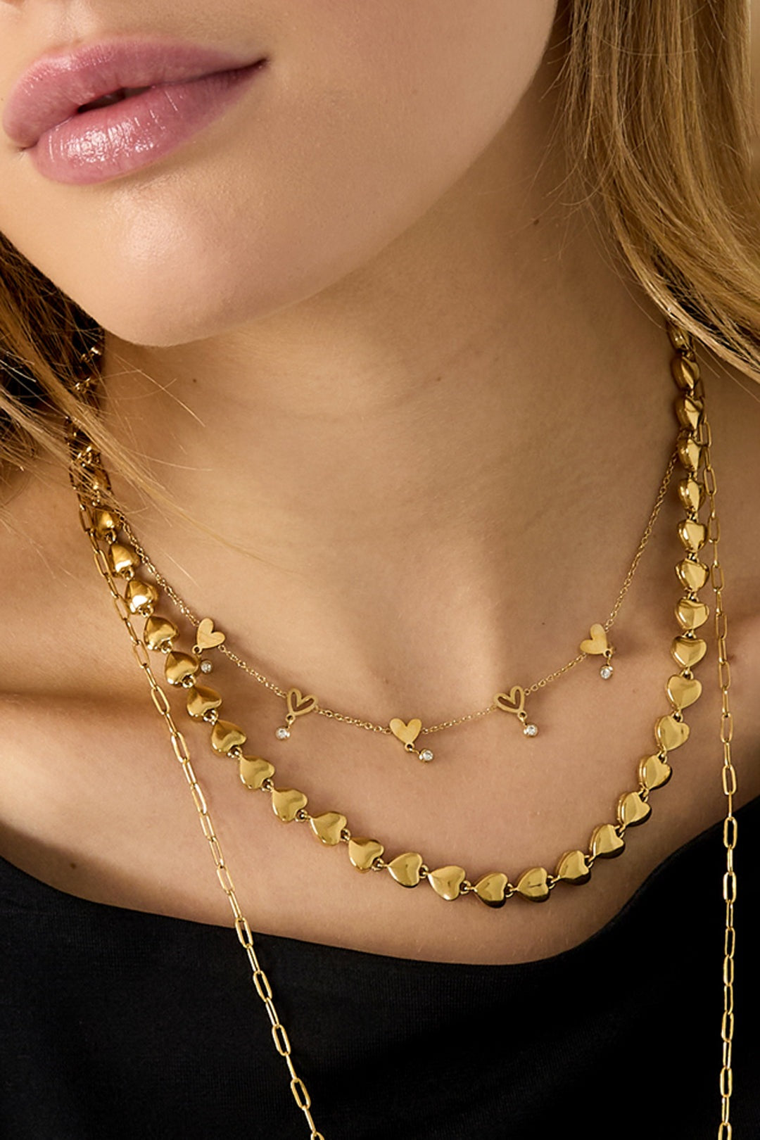 Close-up of model's neck showing detailed gold heart link chain necklace paired with a delicate heart charm necklace over a black top
